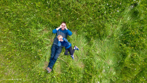 Pappa och son som ligger i gräset, de har gjort händerna till kikare och spanar upp mot himlen. Drönarbild.