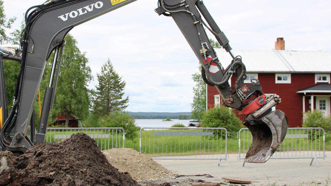 Liten grävmaskin och en jordhög. Rött hus i bakgrunden.