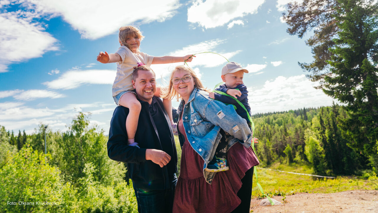 Familj. Pappan har ett barn på axlarna och mamman ett barn på ryggen. Alla är glada.