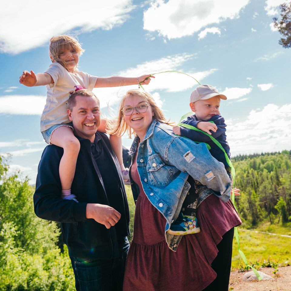 Familj. Pappan har ett barn på axlarna och mamman ett barn på ryggen. Alla är glada.
